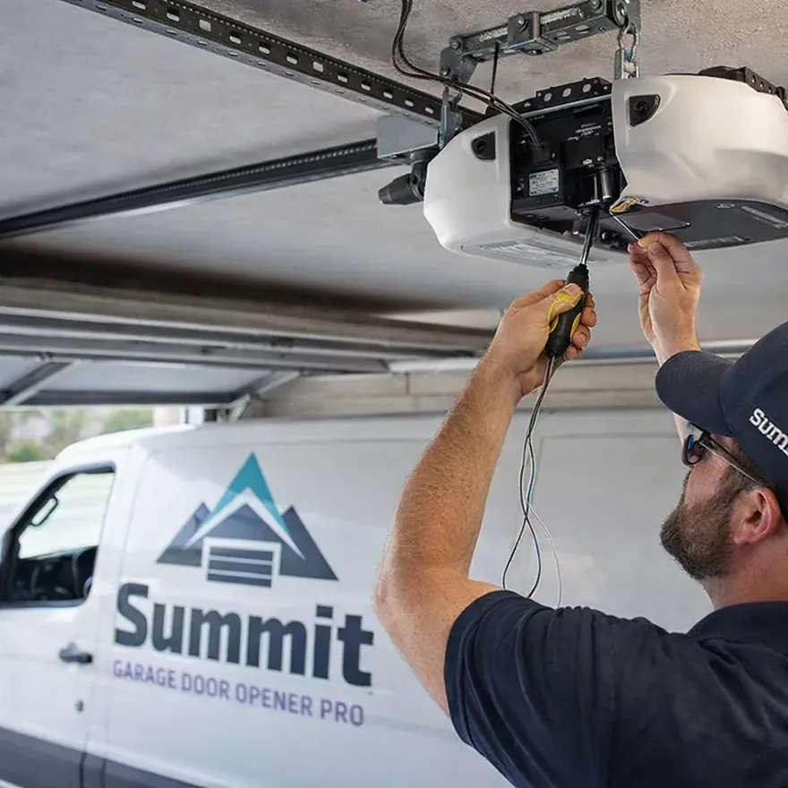 A technician wiring a new garage door opener motor unit mounted to the ceiling, with the rail and trolley visible in a residential garage.